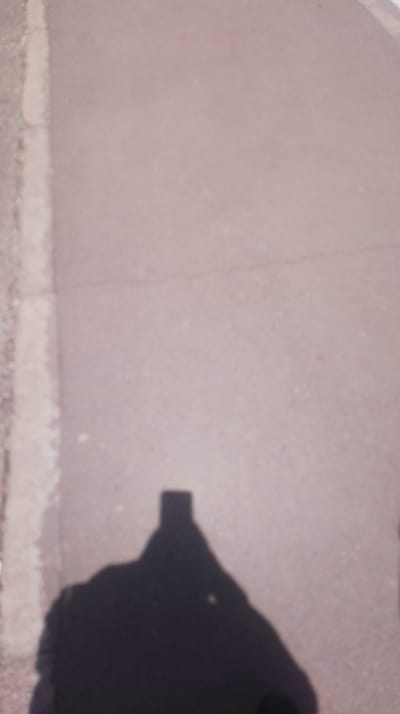 The image displays a ground-level view of a light-toned paved surface, consistent with concrete or asphalt, showing minor surface cracks. A lighter, textured vertical strip, indicative of a curb or pavement edge, occupies the far left of the frame. A prominent, dark human shadow, cast by an overhead light source, extends across the lower central and left areas. Within this shadow, the distinct outline of a rectangular smartphone device held upright is visible, indicating the photo is a self-portrait. No other individuals or significant objects are present. The scene is located in Kikuyu Township ward, Kenya.