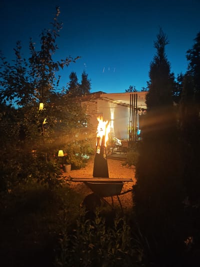 The image depicts a backyard scene at dusk in Надежда Снт, Russia. A tall, narrow metal fire pit is ablaze in the center of the frame, its flames casting a warm orange glow. The fire pit rests on a green wheelbarrow. In the foreground, a leafy tree with three hanging yellow lantern-style lights partially obscures the view. The ground around the fire pit is covered in gravel, and beyond it, a white structure with a covered porch and outdoor seating is visible. Dark silhouettes of trees and bushes frame the scene, with a deep blue twilight sky above. There are no people visible in the image.