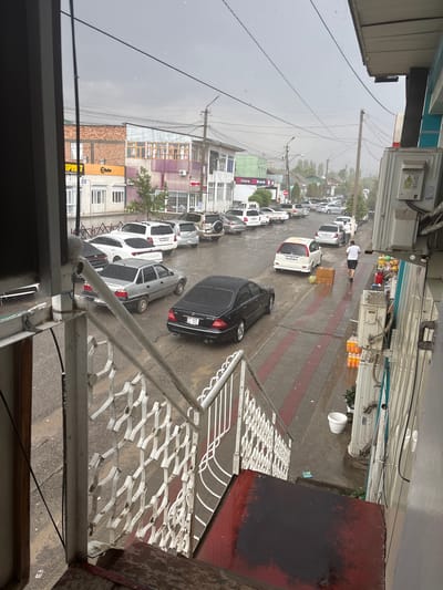 The image depicts a street scene in Batken City, Kyrgyzstan, during what appears to be a rain shower. The perspective is from a high vantage point, looking down and to the side. The street is lined with parked cars, predominantly white SUVs and sedans. A few people are visible, including a person in a white t-shirt and dark shorts walking on the wet pavement.
The setting includes multi-story buildings with commercial storefronts on the ground floors, indicated by signage and window displays. One building has a sign that reads "Store & Bank". The street itself is paved with gray and red tiles, and the wet surface suggests recent or ongoing precipitation. There are power lines crisscrossing above the street.
Notable details include the overcast, gray sky, which points to cloudy or rainy weather, possibly in the late morning or afternoon. The presence of water on the ground and the muted light are consistent with rain. The architecture of the buildings and the types of cars suggest a contemporary urban environment in Central Asia. There is a white bucket and a small green plant near a parked white vehicle. The overall impression is a quiet, everyday scene in a city dealing with inclement weather.