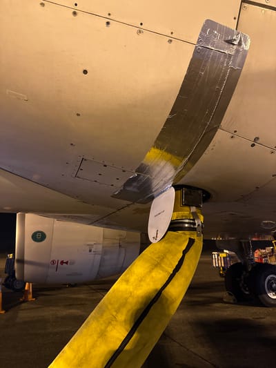 An aerial view of an airplane fuselage shows a large, yellow hose connected to a port on the underside of the aircraft. The hose appears to be an air conditioning or ventilation hose, secured with metal clamps. Silver tape is visible on the fuselage around the connection point, possibly indicating a temporary repair or modification. In the background, the landing gear of the aircraft is visible, along with portions of an engine nacelle featuring a green circular emblem and caution markings. The scene takes place at night on an airport tarmac, indicated by the dark ground, artificial lighting, and the presence of airport equipment. There are no people visible in the image. The location of San Juan Bautista, Peru is not discernible from the image itself.