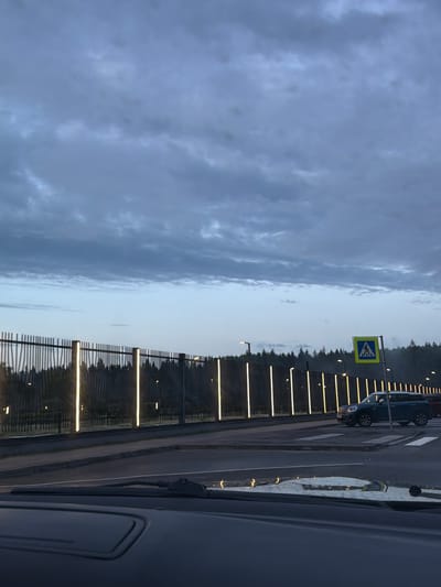 The image shows a twilight view of a road with a pedestrian crossing in Beloostrov, Russia. A dark blue Mini Cooper is stopped at the pedestrian crossing. A yellow pedestrian crossing sign is visible on the right side of the road. A dark metal fence with vertical lights runs along the left side of the road, with a dark forest visible behind it. The sky is cloudy and the overall lighting suggests it is either early morning or late evening. The perspective is from inside a vehicle, with part of the dashboard visible in the foreground.