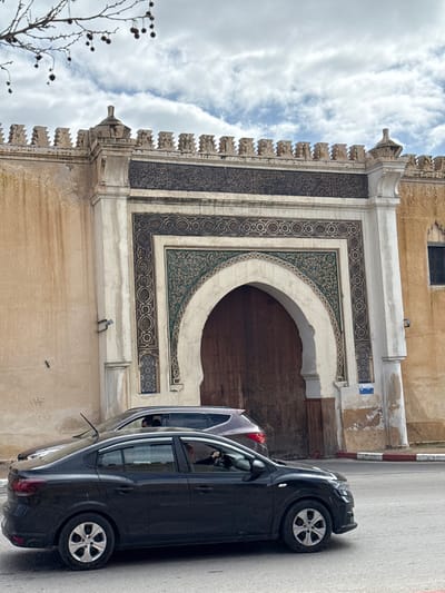 The image displays a street view in Mechouar Fes Jdid, Morocco, centered on an ornate architectural gate. The gate features a large, pointed archway with dark wooden doors, framed by a facade adorned with intricate geometric, arabesque, and calligraphic patterns in shades of green, brown, and off-white. Above the arch, a dark band of Arabic calligraphy is visible, surmounted by a crenellated parapet with a singular finial. The surrounding walls are a light tan color, exhibiting signs of age, staining, and wear, with a small, barred window on the right side. In the foreground, a black four-door sedan is visible, positioned on an asphalt road, partially obscuring the lower section of the gate. A driver's upper torso and hands on the steering wheel are discernible through the sedan's front window. Behind the sedan, a dark grey SUV is partially visible. The road is delineated by a red and white painted curb. Overhead, the sky is overcast with a mixture of white and grey clouds. Bare tree branches with dark, round seed pods or fruits extend into the upper left quadrant of the image.