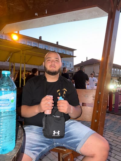 The image shows a young man sitting at a wooden table outdoors, likely at a restaurant or cafe in Debelets, Bulgaria. He is wearing a black t-shirt and denim shorts and has a black Under Armour sling bag across his chest. He is holding a pink can and a bottle of water. In the background, other people are visible, and the setting appears to be a covered outdoor seating area with wooden beams and benches. The lighting suggests it is late afternoon or early evening, with the sky appearing bright. The water bottle has text in Cyrillic, identifying it as "Елинград" (Elinvest), a brand of mineral water. The overall mood seems relaxed and casual, with the man looking directly at the camera with a slight smile.