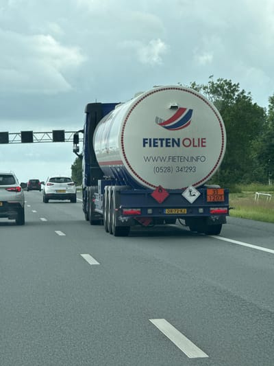 The image shows a tanker truck with the logo "FIETEN OLIE" on its side, driving on a highway. The truck has a large white tank with red trim and a Dutch flag emblem. The license plate of the truck is OR-72-KJ. There are other cars on the highway, as well as trees and a cloudy sky in the background. The scene appears to be captured from a moving vehicle. The location is the A28 highway near Meppel, Netherlands, based on the presence of Dutch license plates and the commonality of this highway for transport in the region.