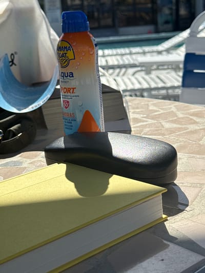 The image presents a static outdoor scene on a patterned light-colored table at a poolside location in Hallandale Beach, United States. In the foreground, a yellow-covered hardcover book with white page edges is visible, partially overlaid by a black, textured glasses case. An orange and blue Banana Boat Aqua Protect Sport SPF 50 sunscreen spray bottle, exhibiting visible water droplets on its label, stands upright behind the glasses case. To the left of the bottle, a portion of a white bag with a blue trim and an 'H' logo is discernible, alongside a dark strap from another item. Direct sunlight illuminates the scene, casting prominent shadows from the objects onto the table and book, including a partial human-like shadow on the book. The background features a clear blue swimming pool, surrounded by multiple white and blue striped lounge chairs. No individuals are directly visible.