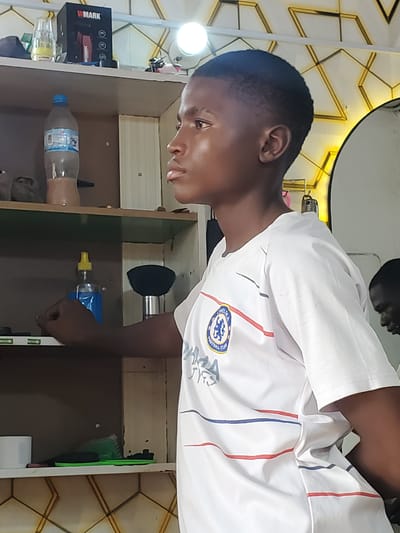 The media file shows a young male subject in what appears to be a barbershop. The subject is wearing a white t-shirt with blue and red stripes and a crest that appears to be for Chelsea Football Club. He is looking down and to the right, with a neutral expression.

The setting includes a shelf unit, which has several items on it. There is a black box with "W MARK" written on it, a bottle of water, and a blue spray bottle with a yellow nozzle. A green barber's tool, possibly a clipper guard or brush, is also visible. The background wall has a geometric pattern with gold accents, and a circular mirror can be seen at the top. The lighting suggests it is daytime. The overall impression is a casual, indoor setting, likely a place of business. No specific weather conditions or emotional states beyond neutrality are discernible.