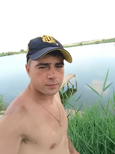 A man wearing a dark blue baseball cap with a yellow emblem and a gold chain necklace stands shirtless in front of a body of water. The man is looking directly at the camera. To his right, there is tall green grass and a wooden dock extending into the water. The background shows a distant shoreline with sparse vegetation and a pale sky. The setting appears to be a natural outdoor environment. There is no indication that the image was taken in Ciumai, Moldova.