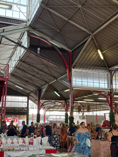 The image displays the interior of a covered market, identified as the Marché de Papeete in Papeete, French Polynesia. The structure features a multi-faceted gray corrugated metal roof, supported by a framework of white and red painted metal beams and columns. Rectangular white translucent panels are integrated into the roof for natural illumination, supplemented by suspended fluorescent tube lights.

Approximately 15-20 individuals, predominantly adults, are present. In the foreground left, a stall displays numerous jewelry items, including necklaces on white stands and various small accessories. Mid-ground stalls exhibit a range of merchandise, such as colorful fabrics, clothing, and carved wooden objects. A woman wearing a blue, off-the-shoulder dress with a swirling pattern stands prominently in the mid-right, holding a light blue bag. Other individuals, comprising both vendors and shoppers, are observed standing, browsing, and interacting around the merchandise displays. Red arched structural elements and green decorative foliage are visible on several columns. The background shows additional market stalls and glimpses of external urban structures through the market's open framework.