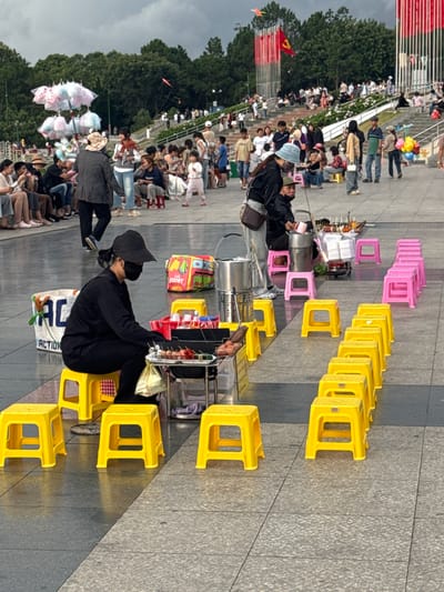 The image depicts a crowd of people gathered outdoors, likely for an event or to observe something. The scene is set on a paved area with tiered seating or steps in the background, populated by numerous individuals. In the foreground and midground, there are many small, brightly colored plastic stools – a mix of yellow and pink – arranged in rows. Several people are seated on these stools, and some appear to be vendors, with carts containing food or goods. One individual in the foreground is wearing black clothing and a black hat, and appears to be a vendor. Another vendor is seen with colorful cotton candy.
In the background, trees and a cloudy sky are visible. A red flag is also prominently displayed, suggesting a possible national or ceremonial event. The people in the crowd are dressed in casual clothing, and their postures suggest they are observing something happening off-camera. The presence of numerous small stools implies a temporary seating arrangement for a gathering. The overall atmosphere appears to be one of public assembly, possibly a festival, market, or a public viewing. The lighting suggests it is daytime, with the sun casting some shadows. There is no readily discernible text in the image, other than a small label on a cart that reads "FOOBY". The location is identified as Da Lat, Vietnam.