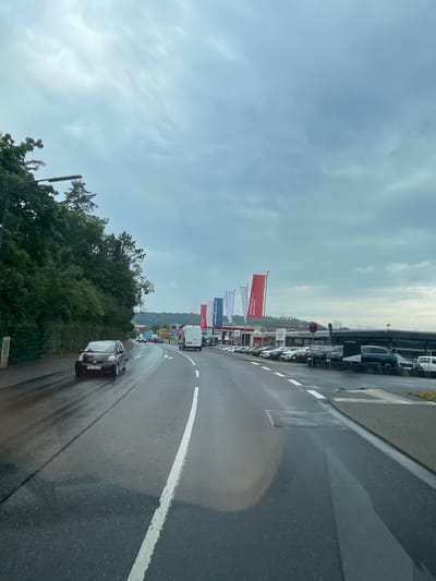 The image depicts a street scene in Lauterbach, Germany, on a cloudy day with wet asphalt, suggesting recent or ongoing rain. The perspective is from a vehicle moving forward on a two-lane road.

On the left side of the road, a dense green embankment covered with trees rises sharply. A black compact car is visible driving in the left lane, its headlights on. To the right of the road, a commercial area is visible. Several cars are parked in rows, likely in a dealership or a parking lot. A row of tall flags, predominantly red and white, are planted in front of the buildings. Some of these flags display text, but it is mostly illegible.

A white van is moving in the right lane of the road. The sky is overcast with thick, gray clouds. The overall impression is of a mundane, everyday scene in a suburban or semi-urban setting in Germany.