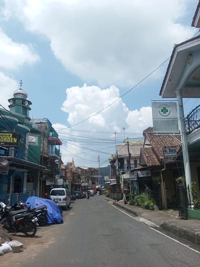 The image depicts a street in a densely populated area, likely a town or city. The road is paved and appears to be mostly empty, with parked cars and motorcycles on the sides. Buildings line both sides of the street, exhibiting a mix of architectural styles, including traditional and modern elements. Some buildings are adorned with signs, including those in Indonesian such as "BAROKAH" and "dr. Dara," suggesting the location may be in Indonesia rather than Laos. There is a mosque tower visible on the left side of the street. Overhead, numerous electrical wires crisscross the sky. The sky itself is bright, with scattered clouds. People are present, although not prominently featured. Overall, the scene portrays a typical urban street view in Southeast Asia.