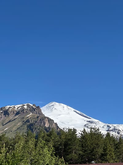 In Terskol, Russia, a photograph captures a clear blue sky above snow-capped mountains. In the foreground, a dense line of dark green evergreen trees forms the bottom third of the image. Behind the trees, the rugged, rocky slopes of a mountain rise, partially covered in patches of snow and green vegetation. Further in the background, a larger, majestic mountain peak, entirely blanketed in snow, dominates the scene. The overall impression is one of natural beauty and grandeur, with the snow-capped mountains serving as the primary subject under a vast expanse of blue sky.