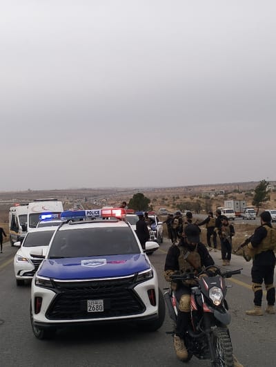 A line of police cars and other vehicles, including an ambulance, is on a road. Several police officers in tactical gear, some holding rifles, are standing beside the vehicles. In the foreground, an officer sits on a motorcycle. The sky is overcast, and the landscape appears arid with distant buildings.