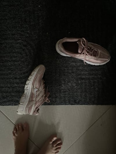 The image shows a pair of pink athletic shoes, one of which is in focus and the other slightly out of focus, resting on a dark textured rug. The rug is bordered by a tiled floor in a lighter color. In the foreground, a person's bare feet are visible on the tiled floor, positioned between the two shoes. The lighting suggests an indoor setting. The location is identified as Novogorelovo, Russia.