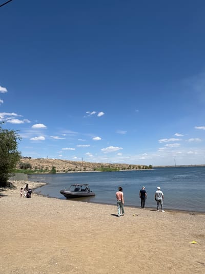 A wide shot captures a tranquil scene on the shore of a body of water, likely a lake or river, under a bright blue sky dotted with fluffy white clouds. The foreground features a sandy beach, with some scattered pebbles and debris near the water's edge.
On the beach, several individuals are present. Near the left side, a woman pushes a stroller while a young child stands beside her. Further down the beach, three individuals are depicted near the water's edge. One person, wearing a pink shirt and light-colored pants, faces away from the camera, looking towards the water. Two other individuals stand slightly further away, also facing the water. One wears a dark shirt and dark pants, and the other wears a light-colored shirt and light-colored pants, with a white cap. A small yellow object, possibly a toy, is visible on the sand near the group of three.
A motorboat with a canopy is moored close to the shore, bobbing gently in the water. The middle ground is dominated by the expanse of the water, reflecting the blue sky.
The background reveals a dry, arid landscape with rolling hills covered in sparse vegetation. A line of trees runs along the edge of the water in the distance. Two power line towers are visible on the hills, and another taller tower stands on the horizon to the right. A fence runs along a portion of the shoreline on the left side of the image. The overall impression is one of a peaceful, sunny day in an outdoor recreational area. This scene is likely located in or around the city of Jezqazğan, Kazakhstan, a region known for its steppes and proximity to water bodies.