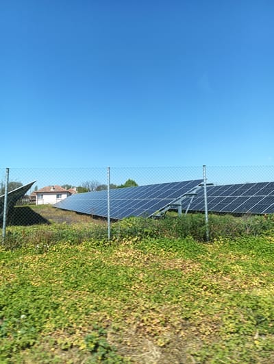The image shows a solar panel field in Straldzha, Bulgaria. The panels are arranged in rows and angled towards the clear blue sky. A chain-link fence surrounds the field. Grass and various green and yellow plants grow in front of the fence. In the background, there is a small house with a reddish-brown roof and white walls.