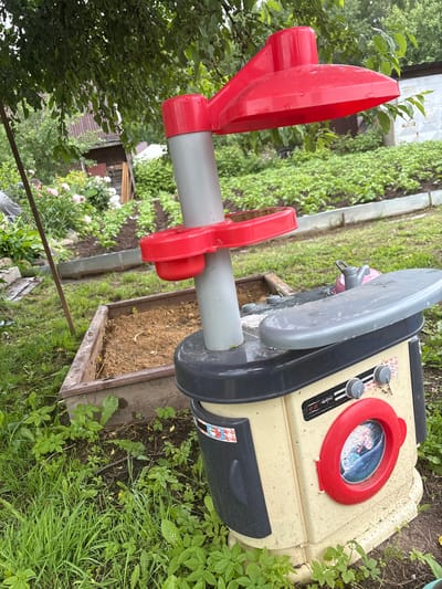 In Golenishchevo, Russia, a weathered plastic toy kitchen is situated outdoors amidst overgrown grass and a garden. The toy, predominantly beige and black with red accents, features a stovetop, oven door, and what appears to be a washing machine compartment. A small pink toy kettle rests on the stovetop. Behind the toy, a wooden raised garden bed is visible, filled with dark soil and a few scattered leaves. Beyond that, rows of green plants stretch across the garden, bordered by metal edging. A rustic wooden shed and a white building are partially visible in the background, nestled amongst trees. The scene is captured under a canopy of green leaves from a nearby tree, suggesting an outdoor domestic setting.
