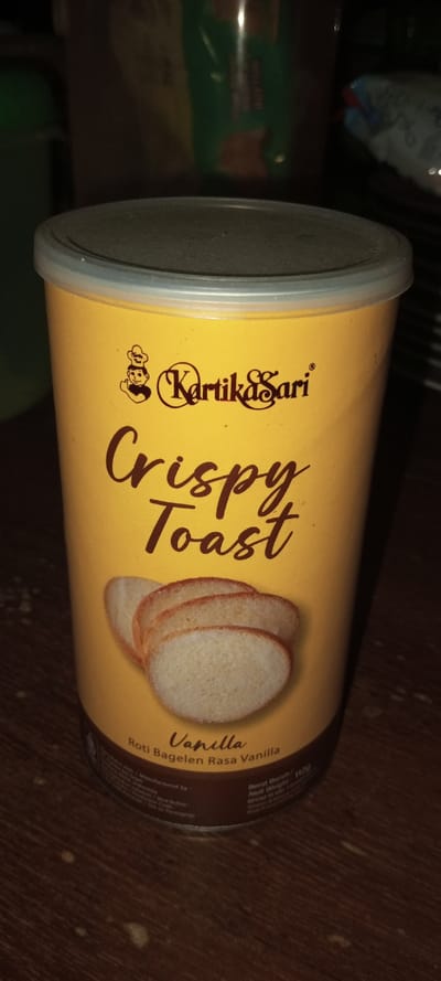 A yellow can of Kartika Sari Crispy Toast is the focal point. The can features a dark brown script font for "Crispy Toast" and an image of sliced toast. The flavor is indicated as Vanilla and “Roti Bagelen Rasa Vanilla”. The can has a plastic lid. The background is dark and cluttered, with hints of other containers and objects. The setting appears to be indoors.