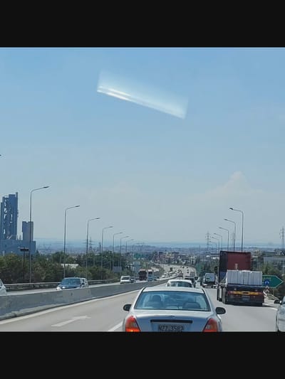This is a photograph taken from a vehicle moving on a highway, likely during the daytime under a clear, bright sky with some glare. In the upper portion of the sky, there is a prominent, elongated, bright white reflection or anomaly. The highway is multi-lane, with concrete barriers separating directions of travel. Several vehicles are visible on the highway, including cars and trucks. One car in the foreground has a license plate that reads "NZ7 3582". In the distance, a city skyline is visible, featuring industrial structures, buildings, and power lines. The landscape appears somewhat arid with some vegetation. Specifically within the city of Bielawska Wieś, Poland, the scene depicts a section of a highway, characterized by its infrastructure and the flow of traffic.