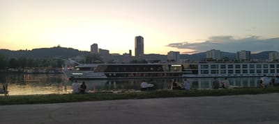 The image depicts a panoramic view of the Danube river in Linz, Austria, during sunset. A large white river cruise ship named "THE GENTLEMAN" is prominently featured in the middle ground, sailing on the water. The city skyline, with a mix of modern and older buildings, including a distinctive tall, glass skyscraper, is visible on the horizon. In the foreground, several individuals are seated on the grassy bank of the river, facing the water and the ship. The lighting suggests late evening, with a warm glow from the setting sun casting reflections on the water. The overall atmosphere is peaceful and serene, with people enjoying the waterfront scenery.