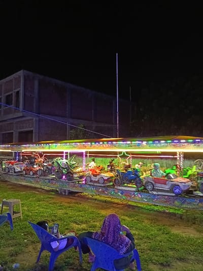 This nighttime image, captured in Namplohbaro, Indonesia, displays an outdoor amusement setting. In the midground, a brightly lit children's carousel-style ride operates, featuring a circular track with various miniature vehicles, including cars, motorcycles, and ATVs. Several children, indistinct in detail, occupy the vehicle seats. The ride's structure is adorned with multi-colored LED lights, creating a vibrant illumination effect. In the foreground, two individuals are seated on blue plastic chairs on a patch of short green grass. One person, seen from behind, wears a purple patterned head covering and faces towards the ride. The other individual, partially visible to the left, holds and observes a mobile phone. The background consists of a dark sky above a multi-story, unfinished brick building with exposed concrete frames. A tall, slender white pole stands vertically near the center-right of the background, with dark foliage visible beyond.