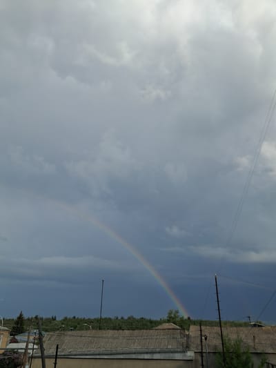 The image depicts a cloudy sky with a visible rainbow arching across it. In the foreground, there are rooftops of houses, some with satellite dishes and electrical wires. A line of trees is visible in the middle ground, and in the distance, the sky darkens, suggesting a storm. The scene appears to be an outdoor view from a residential area. The location is identified as Gavar, Armenia, implying this is a view of the sky and cityscape in that region.