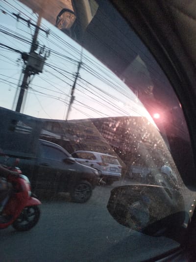 The image, taken from inside a vehicle, shows a street scene in Tanjungkarang, Indonesia. In the foreground, a red scooter is partially visible on the left, with its front wheel and part of its body in frame. To the right of the scooter, a dark-colored car is seen from its side. In the background, a light-colored SUV is parked or moving slowly. Above the vehicles, numerous electrical wires crisscross the frame, supported by utility poles. The sky is visible above the wires, with a bright lens flare from the sun reflecting off the car window. The overall scene suggests a typical urban environment in Indonesia with traffic and infrastructure.