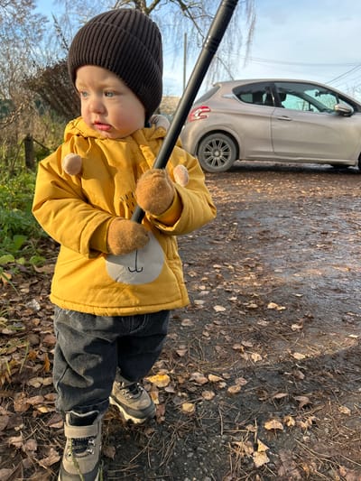 A child in a dark beanie, yellow coat, grey jeans, and hiking boots stands in a grassy area with fallen leaves. The child holds a black pole with both hands. A light brown car is parked on the right side of the frame. The ground appears wet with a mix of grass, leaves, and dirt. The sky is cloudy in the background.