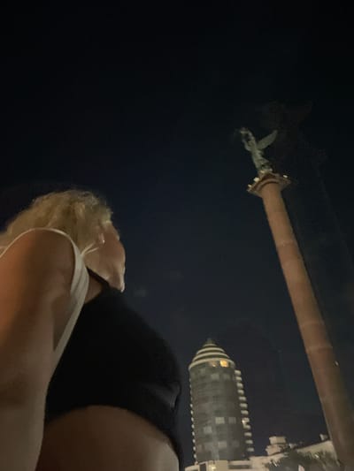 The image, taken at night, features a woman with blonde hair in the foreground, looking towards a monument in the distance. The monument is a tall column topped with a statue of a winged figure. Behind the monument, a modern, circular building with illuminated windows is visible. The scene appears to be set in an urban environment. The location is Merchanskoe, Russia.
