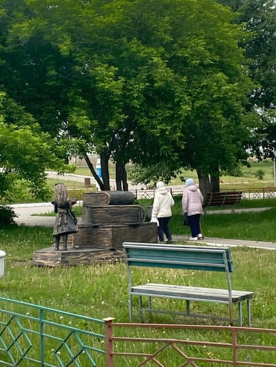 In Belorechensky, Russia, a statue of a young girl stands next to a sculpture of stacked books in a grassy area. Two elderly women, one in a white jacket and the other in a pink jacket and blue hat, walk past the sculpture. A park bench is situated in the foreground, and a fence runs along the edge of the park. Trees and a pathway are visible in the background.