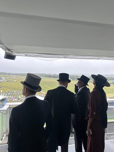 Four individuals, three men and one woman, are observed from behind on a covered outdoor viewing platform, likely at Epsom Downs Racecourse in Epsom and Ewell, United Kingdom. The sky is overcast, and wet surfaces on the platform and railing indicate rain or recent precipitation.

On the far left, a man wearing a grey top hat and a dark suit looks out over the scene; an earring is visible on his left ear. To his right, a second man in a black top hat and dark suit also looks forward. Adjacent to him, a third man in a black top hat, dark suit, and glasses holds his right hand to his ear, possibly using a communication device or phone. On the far right, a woman in a wide-brimmed black hat and a dark reddish-brown, long-sleeved dress holds a white object, likely a smartphone, in her left hand as she gazes slightly upwards and to the right.

Above them, a light-colored flat ceiling is visible, with a black rectangular speaker or monitor mounted to it on the left. A metal railing with transparent panels defines the edge of the viewing platform. Beyond the platform, a vast green grass racetrack stretches into the distance under the uniformly grey sky. To the left of the track, a large parking area is filled with numerous parked cars, alongside some blue and white event structures. A dense line of trees forms the distant horizon.
