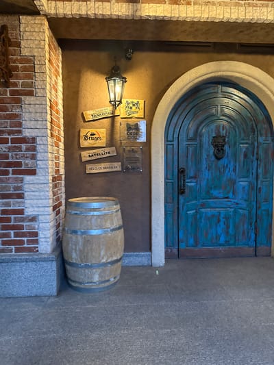 This is a photograph of a rustic entrance with a distressed blue door, an arched stone surround, and a brick facade. The door has a dark metal handle and a decorative knocker. To the left of the door, a gas lamp illuminates a section of the wall. Several wooden signs with various logos and text are mounted on the wall. One sign reads "TANKBRAU" and another "SATELLITE". A large wooden barrel rests on a gray stone surface to the right of the signs. The overall setting suggests a pub, brewery, or an establishment with a historical or European theme. The lighting indicates it might be evening or indoors. No people are visible in the image. The context of Oktyabr'skoe, Russia, is provided as location information.