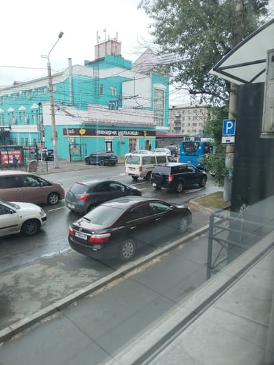 A street scene in Chita, Russia, shows a turquoise building with multiple windows and architectural details, possibly a commercial establishment given the signage that reads "Пекарня" (Bakery). Several vehicles are present on the wet asphalt road, indicating recent rain. These include a dark sedan in the foreground with a license plate reading "P 300 HK 75," a gray hatchback behind it, a white sedan, a brown minivan, a black SUV, and a blue bus with the number 352 visible. A parking sign with a "P" symbol is also visible on a pole. The environment appears to be urban, with overhead wires, streetlights, and other buildings in the background. The sky is overcast.