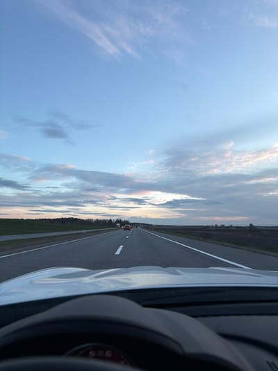 The image captures a view from inside a vehicle driving on a highway or rural road, likely during either dawn or dusk. The sky, a dominant feature, occupies the upper portion of the frame, displaying gradients of light blue merging into warmer hues of pink and orange near the horizon, indicative of the low sun. Scattered, wispy clouds are visible across the sky.
A long, straight asphalt road extends from the foreground into the distance, marked by prominent white lines that divide the lanes. On the right side of the road, a flat, green landscape with a distant line of trees borders the asphalt. Further along the road, several vehicles can be seen, appearing as small silhouettes in the distance.
The interior of the vehicle taking the photo is partially visible in the foreground, showing the white bodywork on the left, and elements of a dark dashboard and steering wheel rim on the right. A small, circular red gauge with white markings, possibly reading "S=6" in a blurred font, is faintly discernible on the dashboard. The scene suggests a tranquil journey through an open landscape, consistent with a rural area like Пекалінскі сельскі Савет, Belarus.