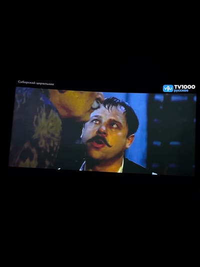The image shows a close-up of a man's face with a prominent mustache, looking upwards with his mouth open. He appears to be in distress or surprise. The background is dark and blurry, with some indistinct shapes and colors. In the top left corner, there is text in Russian that reads "Сибирский цирюльник" which translates to "The Siberian Barber". In the top right corner, there is a logo for "vju TV1000 русское". The scene is likely from a movie or television program. There is no visible indication of the city of Kokkorevo, Russia in the image.