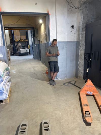 A man in a grey t-shirt and dark shorts stands in a doorway, looking at his phone. He is in a warehouse with concrete floors and walls, with a forklift visible in the background. An orange pallet jack is in the foreground. The scene appears to be in Djerman, Bulgaria.