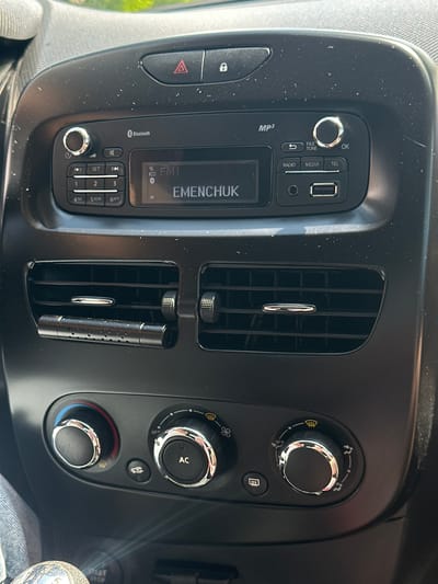 The image shows the interior of a car, specifically the dashboard area. A car's audio system with a display screen is prominently featured, showing "FM1" and "EMENCHUK". There are various buttons and dials for controlling the radio, media, and phone functions. Below the audio system are two air vents, and further down are climate control dials, one of which is labeled "AC". The dashboard appears dusty. The surrounding environment outside the car is not visible. The image was taken in Mala Kokhnivka, Ukraine.