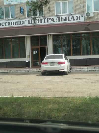 The image captures the facade of a brick building, likely a hotel given the signage that reads "Гостиница 'Центральная'" (Hotel 'Centralnaya'). The building features large windows with dark frames and an entrance with glass doors. A white sedan is parked in front of the building, partially obscuring the lower portion of the entrance. An air conditioning unit is visible on the upper right side of the building. The area in front of the building appears to be a dirt or gravel lot, with a strip of green grass bordering it. The location is identified as Lesozavodsk, Russia.