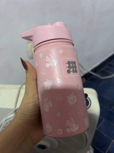 A hand holds a pink water bottle, decorated with cartoon characters, against a background of a white fan and blue-tiled wall. The location is not discernible from the image.