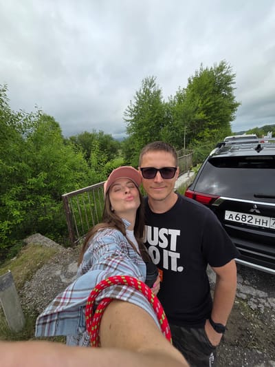 A selfie taken outdoors shows a man and a woman standing next to a black Mitsubishi Outlander. The woman is on the left, wearing a pink baseball cap, a blue and white plaid shirt over a white t-shirt, and her arm is holding the phone. She is puckering her lips towards the camera. The man is on the right, wearing a black t-shirt with "JUST DO IT." printed in white, sunglasses, and camouflage shorts. He has his hand in his pocket. Behind them is lush green foliage, a railing, and a cloudy sky. The license plate of the car reads "A682HH 7". The environment appears to be a roadside with a natural, somewhat undeveloped setting, likely on the outskirts of a town or village. The city of Теплоозёрск, Russia, is a possibility for the location, given the car's license plate region.