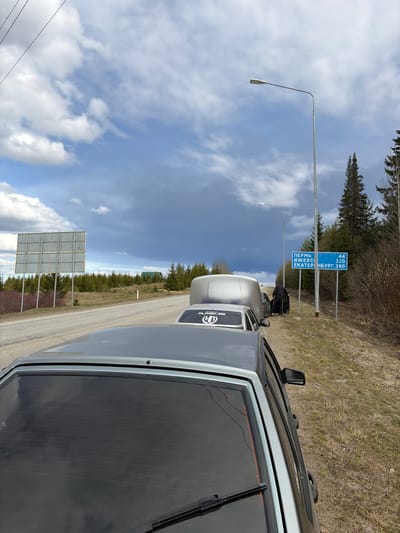 The image shows a section of highway in Yugo-Kamskiy, Russia. Visible are the edge of a silver car in the foreground, two other parked cars, a roadside sign indicating distances to Perm (44 km), Izhevsk (320 km), and Yekaterinburg (390 km), a streetlight, and various road signs. The sky is partially cloudy. The environment includes trees along the roadside. A person is seen walking with luggage.