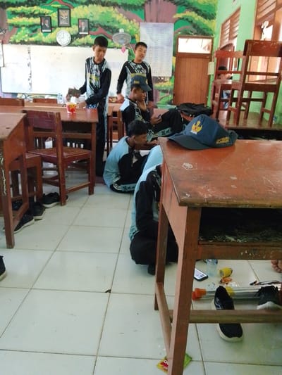 The image depicts a classroom in Sumber Tlaseh, Indonesia, with several male students present. In the foreground, a wooden desk is prominent, with a blue cap bearing Indonesian text ("SMA NEGERI 3 BOJONEGORO") placed on it. To the left of this desk, another wooden desk and chair are partially visible. A student wearing a light blue long-sleeved shirt and dark pants is seated on the floor in front of the desk, looking down. Further back, two students in matching black and blue long-sleeved shirts and dark pants are standing near a whiteboard. One of them is holding a red cup. To their right, another student in a similar uniform is also standing. A third student is seated on the floor behind the first seated student, looking at a mobile phone. The room has tiled flooring and the walls are decorated with paintings. Wooden chairs and desks are scattered throughout the space. The overall scene suggests a classroom setting during a casual moment.