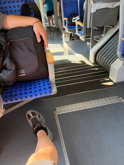 The image depicts the interior of a train carriage, likely in Lüdinghausen, Germany, showing a section of seating and the aisle. In the foreground, a person's legs and feet are visible, with one foot clad in a grey and pink athletic shoe. The person appears to be seated, with a portion of a black backpack resting on the blue and black checkered seat beside them. The backpack has an orange zipper detail and a small brown tag. A dark grey jacket is draped over the backpack and the seat. The flooring of the train is a speckled grey material, with a metal diamond plate strip running along the edge of the aisle. Further into the carriage, another seat is visible with a black Nike drawstring bag on it. Sunlight streams in from the right, casting shadows across the floor and highlighting the ribbed metal stairs and partitions of the train's structure. In the background, partially obscured, are other passengers, including one wearing a bright turquoise t-shirt and dark shorts, and another individual standing. The overall impression is of a typical train journey.
