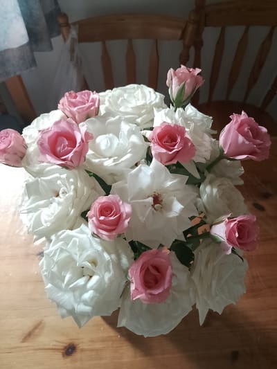 A close-up, overhead shot depicts a bouquet of white and pink roses arranged in a vase, placed on a wooden table. The roses are the central focus, with their petals varying in fullness and stage of bloom. Some white roses are fully open, revealing their intricate layers, while others are more tightly budded. The pink roses are scattered throughout the bouquet, with a few more prominent than others. The wooden table's grain is visible, adding a natural texture to the scene. In the background, a wooden chair with a curved back is partially visible, suggesting an indoor setting. The lighting appears to be natural, casting soft shadows and highlighting the delicate features of the flowers. The image does not provide any specific indicators of its location within Rheda-Wiedenbrück, Germany.