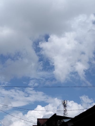 A partly cloudy sky fills the frame, revealing patches of blue. Beneath the sky, a set of parallel power lines stretches horizontally. A telecommunications tower rises above the roofs of buildings below. Dark rooftops contrast with the bright sky, and the tower appears to be surrounded by white, puffy clouds.
