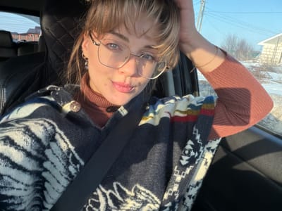 A young female with blonde hair, styled with bangs, is seated in the front passenger seat of a vehicle in Азьмушкино, Russia. She wears large, clear-framed eyeglasses, a rust-colored turtleneck sweater, and a patterned dark blue, white, red, yellow, and light blue poncho or shawl, secured by a black seatbelt. Her left ear displays hoop earrings, one with a dangling dark bead. A thin gold chain bracelet is visible on her right wrist, as her right hand is positioned at the back of her head. She is looking forward, with slightly parted lips. Strong sunlight illuminates the left side of her face and the car's interior. The vehicle's interior features dark upholstery, including a diamond-stitched seat cover on the driver's seat. Through the windows, a winter or early spring landscape is visible under a clear blue sky, featuring patches of snow, bare trees, utility poles with lines, and distant residential buildings, one of which is light-colored.