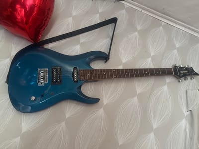 The image shows a blue electric guitar hanging on a wall with white floral patterned wallpaper. A red heart-shaped balloon is partially visible in the top left corner. A black guitar strap is attached to the guitar and extends upwards. The guitar has a dark fretboard with white fret markers and appears to have two pickups. The headstock of the guitar is visible on the right, with tuning pegs and the brand name "Zombie" printed on it. The lighting suggests an indoor environment. There is no visible indication of the location being in Разведчик, Russia.