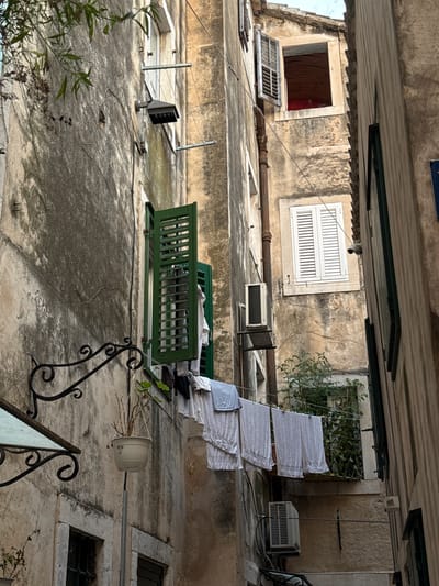 The image depicts an outdoor scene under an overhang, likely in Split, Croatia. The architecture suggests an older building with weathered stone walls and corrugated roofing. In the foreground, a worn concrete surface is visible, with various objects scattered around. A green wooden bench sits prominently in the middle ground, appearing to be assembled from sections. To the left of the bench, a modern, dark gray outdoor light fixture is mounted on a pole. Several electrical wires are strung across the scene, connecting to different devices and possibly lighting.
Above the bench, laundry or fabric sheets are hung out to dry on a line, creating a cascade of white material against the rough wall. A small, green plant in a pot hangs from a metal bar on the right side of the image, its branches bare and dry. Further up the wall, two windows are visible. The left window is open, revealing a dark interior, while the right window is a traditional slatted design. On the upper right, an air conditioning unit is mounted on the wall, suggesting a warmer climate.
The overall impression is one of a lived-in, somewhat cluttered space, possibly a residential courtyard or balcony. The lighting suggests it is daytime, but the overcast conditions and shadows create a somewhat muted atmosphere. There is no visible text in the image.
