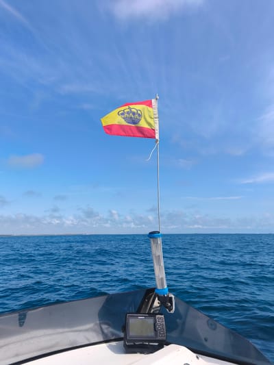 The image captures a forward-facing perspective from the bow of a white recreational boat, navigating open blue waters, specifically located off the coast near the city of Sueca, Spain. In the immediate foreground, the white fiberglass hull of the boat is visible, overlaid by a dark, tinted plexiglass windshield. Mounted on the dashboard is a Garmin GPS/fishfinder unit, displaying operational data. A thin metallic pole extends upwards from the bow, secured near the windshield. Attached to this pole, below the flag, is a transparent cylindrical object with blue banding, possibly a mast light or container. A small Spanish flag, featuring a red and yellow horizontal stripe design with a central gold crown emblem, is affixed to the pole and unfurls in the breeze. The midground is dominated by the deep blue expanse of the sea, exhibiting a gently rippled surface indicative of mild conditions. Along the distant horizon, a low-lying landmass is faintly visible, with indistinct structures or buildings appearing on its edge. The sky above is a clear, bright blue, interspersed with sparse, wispy white clouds. No individuals are visible in the scene.