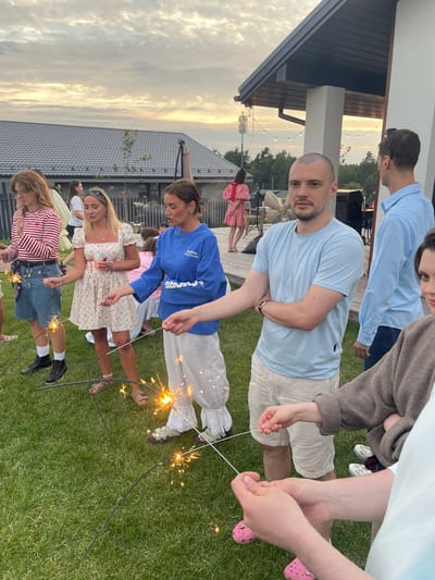 In Simagino, Russia, a group of young adults are gathered on a grassy lawn in front of a modern house. The time appears to be dusk, with soft light illuminating the scene and the sky filled with clouds. Several individuals are holding and igniting sparklers, creating trails of light and sparks. The people are dressed casually, with one woman in a floral dress, another in a blue sweatshirt and white pants, and a man in a light blue t-shirt and beige shorts. The house features a dark roof and white exterior walls, with string lights visible on the porch. The overall atmosphere suggests a festive gathering.